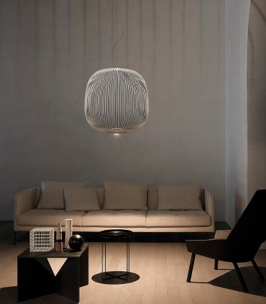 Et minimalistisk stue med Spokes pendellampe fra Foscarini som elegant midtpunkt. Lampens filigrane, metalliske stivere kaster bløde lysspil på væg og loft og giver rummet en let, skulpturel effekt. Lys sofa, mørke accenter og naturlige materialer skaber en rolig, moderne atmosfære