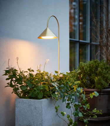 Lysende udendørslampe fra Frandsen ved siden af et beplantet betonkar på en terrasse foran en moderne husfacade.