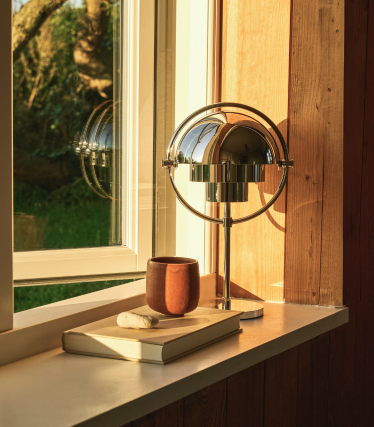 Forkromet bordlampe i en vindueskarm i aftenlyset, ved siden af en bog og et keramikkrus.