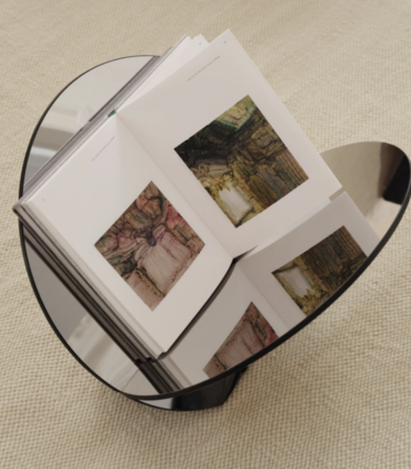 Rundt sidebord i mørkt glas med en åben kunstbog på et beige tæppe.