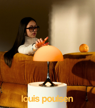 Kvinde med solbriller rører ved en orange Louis Poulsen bordlampe fra den nye Panthella-kollektion. Lampen står på et hvidt sidebord foran en orangefarvet sofa i et varmt, blødt lys.